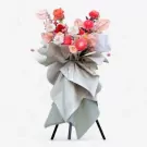A grand floral stand with vibrant coral and peach flowers, wrapped in pale grey paper, on a white background.