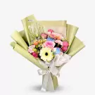 A vibrant bouquet of pink roses, gerberas, blue carnations, and yellow blooms wrapped in light green paper with a white ribbon.