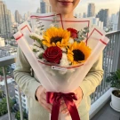 Person holding a bouquet of sunflowers, red roses, and white filler flowers, tied with a burgundy ribbon.
