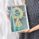 White jasmine malai with blue ribbon in a white box, held by a person in a navy patterned dress.