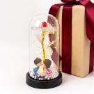 Glass dome with holographic rose, kissing couple figurines, and fairy lights next to a wrapped gift.