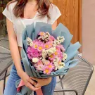 Pink and purple mums with white statice in blue paper wrap, held by a person wearing a white top and jeans.