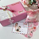 Pink gift box with ribbon, rose candle in clear box, and Valentine's Day cards and stickers.