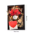 A framed gift box with red roses, a teddy bear, and a card, illuminated by fairy lights.