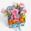Fruit basket with pink roses, blue hydrangeas, oranges, kiwis, and apples, tied with a blue ribbon.