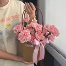 Pink carnations arranged in a brown bag with a pink ribbon, held by a person wearing a t-shirt.