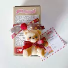 Teddy bear candle with red ribbon, set in a box with Valentine's Day decor and a postcard.
