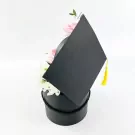 Black graduation hat box with pink roses and white mums, topped with a yellow tassel.