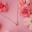 Gold necklace with heart-shaped crystal and bow pendant on a pink background with flowers.