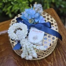 Blue artificial garland and flowers in a bamboo basket, with a blue ribbon and small card.