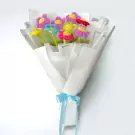 Bouquet of colorful plush daisy flowers with smiling faces, wrapped in white paper and tied with a blue ribbon.