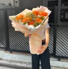 A bouquet of orange roses, alstroemerias, and daisies wrapped in light pink paper.