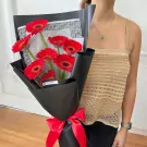 Red gerbera daisy bouquet wrapped in black paper, tied with a red ribbon, held by a person wearing a tan top.