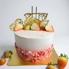 A round, pink and white tres leches cake topped with sliced strawberries and a "Happy Birthday" topper.