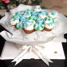Cupcake bouquet with blue, white, and green frosting in brown wrappers, presented in white tissue paper and a white box with ribbon.