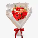 A bouquet of red gerbera daisies, roses, and carnations wrapped in white paper, tied with a red ribbon.