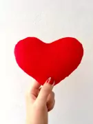 Hand holding a red heart pillow against a white wall, with a heart design on red nail polish.