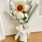 Crocheted bouquet with a sunflower, tulip, daisies, and carnations, wrapped in pale green paper with a cream ribbon.