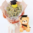 A graduation bouquet of white daisies with a yellow pom-pom wearing a graduation cap, next to a teddy bear in a graduation cap holding a diploma.