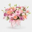 Pink and peach roses, pink gerbera daisies, and purple mums in a white vase with a pink bow.