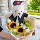 Graduation teddy bear in cap and gown with sunflowers and assorted flowers.