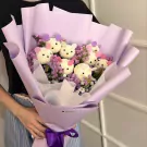 A bouquet of fluffy white teddy bear heads and purple flowers wrapped in lavender paper.