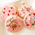 Four pink frosted sugar cookies in a box, decorated with sprinkles and heart shapes.