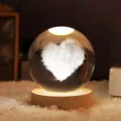 Crystal ball lamp with a 3D carved heart inside, on a wooden base, illuminated with warm LED light.