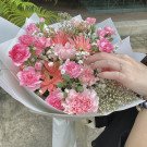 A hand with a diamond ring touches a bouquet of pink carnations, gerbera daisies, and baby's breath.