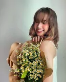Woman smiling, holding a bouquet of white daisies wrapped in brown paper.