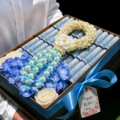 luxury golden gift box featuring 30 neatly rolled banknotes, paired with a spiraled Crown Flower garland and fresh blue blooms