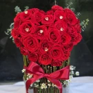 Heart-shaped arrangement of red roses with heart crystals in an acrylic cylinder with a red ribbon.