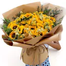 A large bouquet of sunflowers, accented with small white flowers and greenery, wrapped in brown paper.