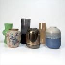 Six vases of different shapes, colors, and materials arranged on a white surface.