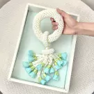 A white beaded jasmine malai with light blue flowers is held in a hand above a white frame.