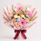 A vibrant bouquet of pink gerberas, roses, tulips, and small white flowers wrapped in pink paper with a burgundy ribbon.