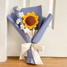 Crocheted sunflower bouquet in blue paper wrap with white flowers and ivory ribbon.