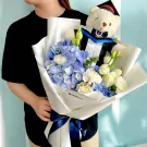 Graduation bouquet with blue hydrangeas, white carnations, and a teddy bear in a graduation gown.