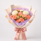A bouquet of white chrysanthemums, peach carnations, and daisies, wrapped in pale cream paper with a pink ribbon.