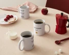 Mugs with "Good Morning Ploy" and "No Speaking" text sit on a table with cherries, cookies, milk, and a red moka pot.