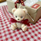Cream teddy bear candle with red ribbon sits on red and white checkered cloth, with gift box in background.