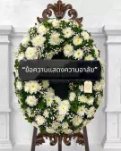 White chrysanthemum wreath with "Condolence message" ribbon, on a dark ornate stand.