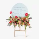 Floral stand with red anthuriums, pink roses, and green foliage on a light blue oval plaque congratulating "cubic EmSphere."