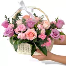 White wicker basket overflowing with pink, purple, and blush roses, carnations, and greenery.