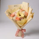 A bouquet of pastel peach roses, white carnations, and a yellow gerbera daisy wrapped in pale yellow paper, tied with a pink ribbon.