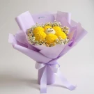 A bouquet of yellow mums arranged in a smile, with white filler flowers and pale purple wrapping.