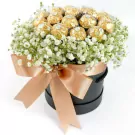 Round black box with white flowers and gold-wrapped Ferrero Rocher chocolates, tied with a rose gold bow.