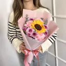 A woman holds a bouquet featuring a bright yellow sunflower, pink carnations, and purple chrysanthemums.