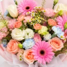 Bouquet of pink and peach gerberas, roses, white tulips, and small white daisies with light pink and white paper wrapping.
