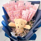 A teddy bear peeks out from a bouquet of Thai 100 baht banknotes and dried purple flowers.